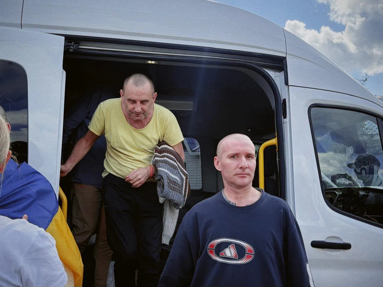 Один із звільнених військових виходить з мікроавтобуса, у руках тримає теплий светр, поруч розгорнутий український прапор