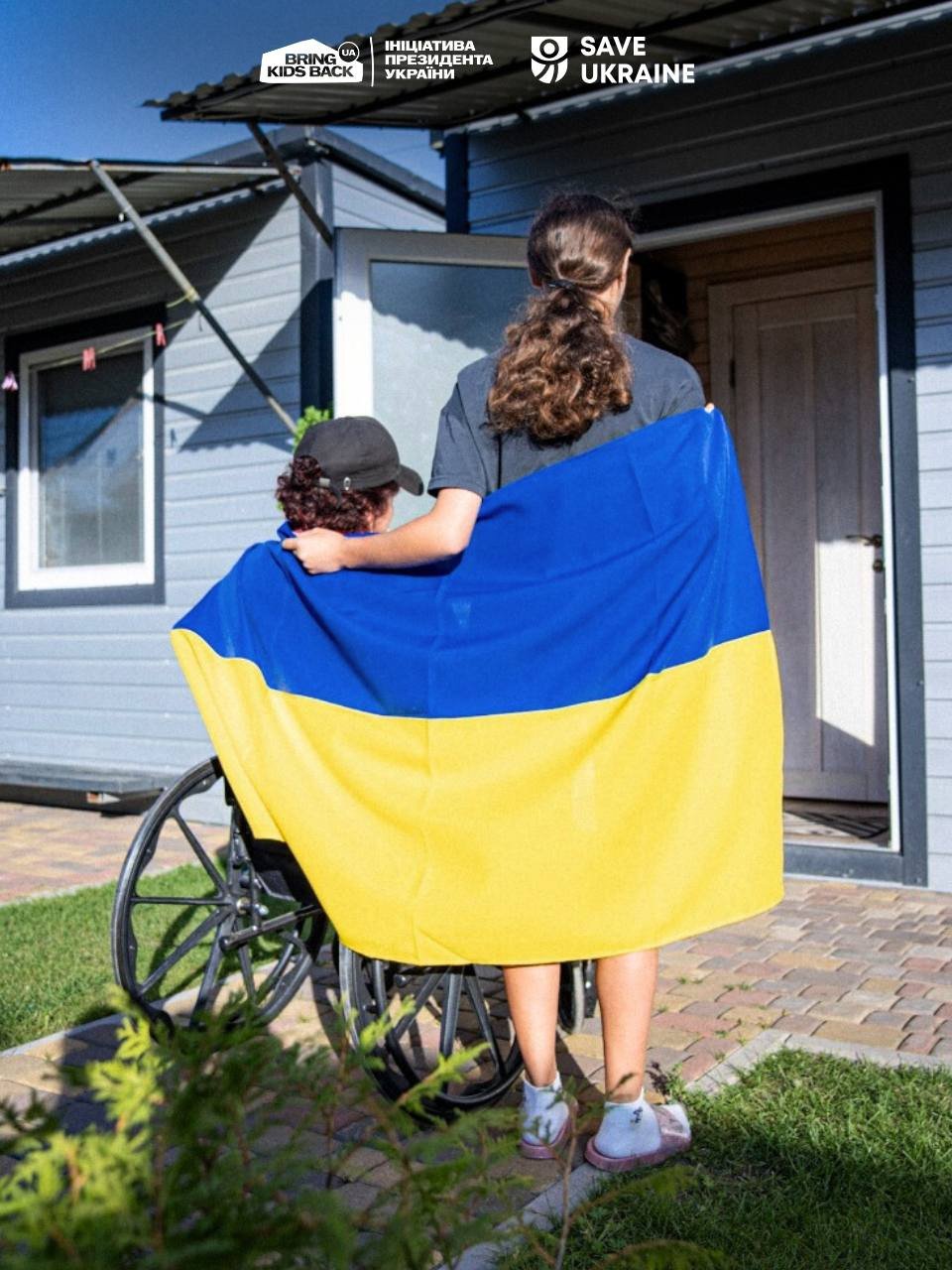 Людина в інвалідному візку і волонтер, загорнуті у прапор України, біля будинку